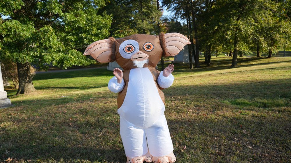A boy, Santiago Hidalgo, is dressed in a large brown-and-white inflatable Gremlin costume with big ears and orange eyes. He stands on a grassy field with trees in the background, raising both hands near his face. He is outside and it is daytime.