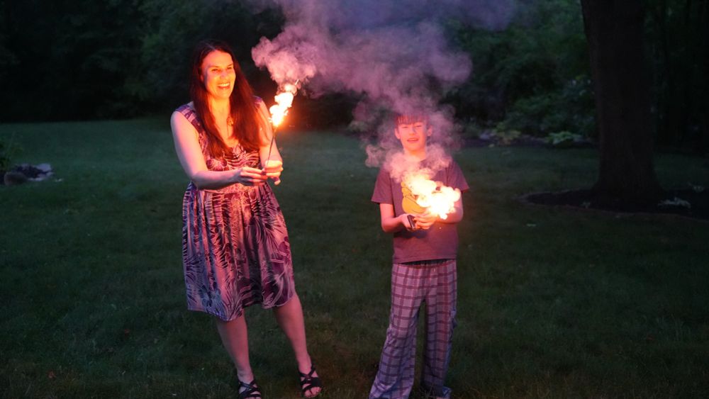 A woman, Alexandra Hidalgo, and a child, Santiago Hidalgo Bowler, hold an orangey-yellow sparkler out in front of them, looking straight at it. They are both outside, with grass and trees behind them. It is nighttime.