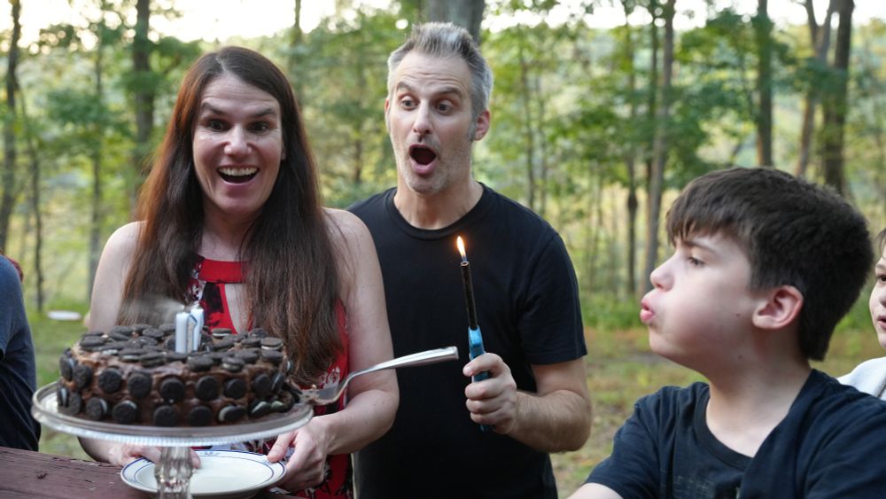 Two adults, Alexandra Hidalgo and Nathaniel Bowler, look at a cake with wide eyes and mouths agape as Santiago Hidalgo Bowler blows out the candles on the top. They are outside, and it is daytime.