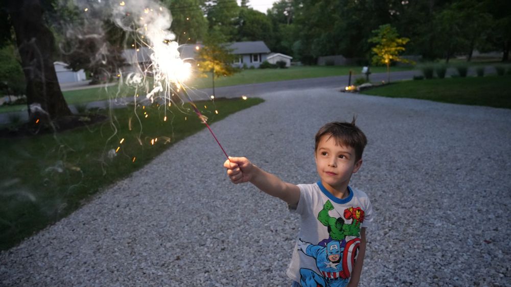 A child stands and holds a sparkler, lit up bright white, facing up on a driveway. He is looking up at the sparkler with a smile. There are trees and a house behind him. He is outside and it is nighttime.