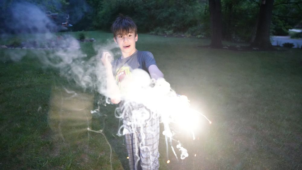 A child stands and holds a sparkler, lit up bright white, facing down in a field of grass. He is looking down at the sparkler in awe. There are trees behind him. He is outside and it is nighttime.