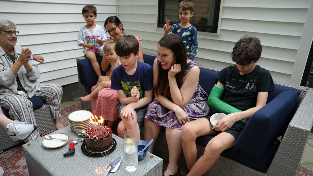 Two children (Santiago Hidalgo-Bowler and William Hidalgo-Bowler) and one woman (Alexandra Hidalgo) sit on a blue couch. Alex is smiling and Santiago is looking down at a chocolate birthday cake on the table in front of them. There are two women and three children beside them, smiling. They are outside and it is daytime.