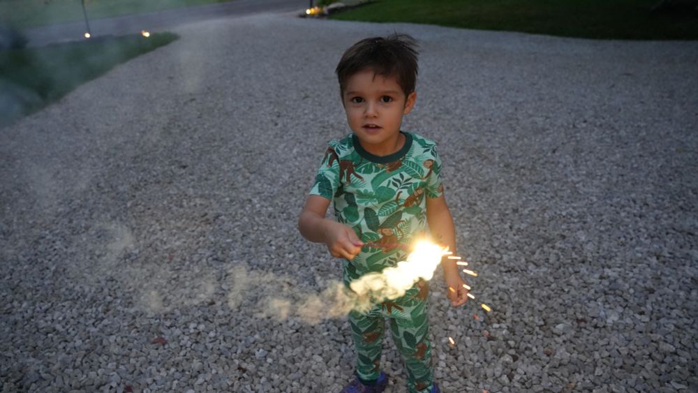 A child stands and holds a sparkler facing down on a driveway. He is outside and it is nighttime.