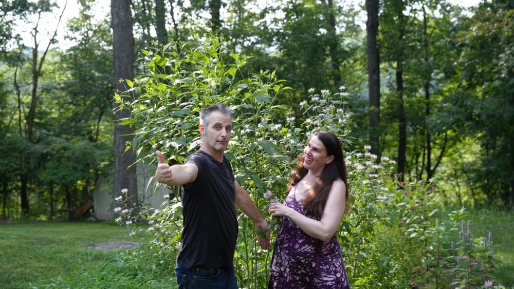 A man (Nathaniel Bowler) and a woman (Alexandra Hidalgo) stand and pose in front of a native garden. Nate is pointing at the camera with his flattened hand and Alex is looking at him with a smile. There are trees behind them. They are outside and it is daytime.