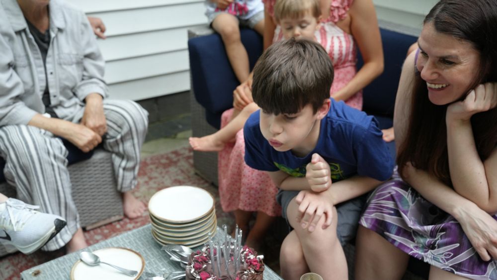 One child (Santiago Hidalgo-Bowler) and one woman (Alexandra Hidalgo) sit on a blue couch. Santiago blows out the candles on his chocolate birthday cake on the table in front of them and Alex smiles. There are two women and three children beside them, smiling. They are outside and it is daytime.