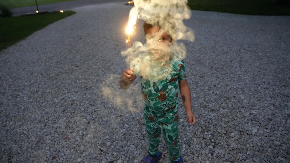 A child stands and holds a sparkler facing up on a driveway, smoke obscuring his face. He is outside and it is nighttime.