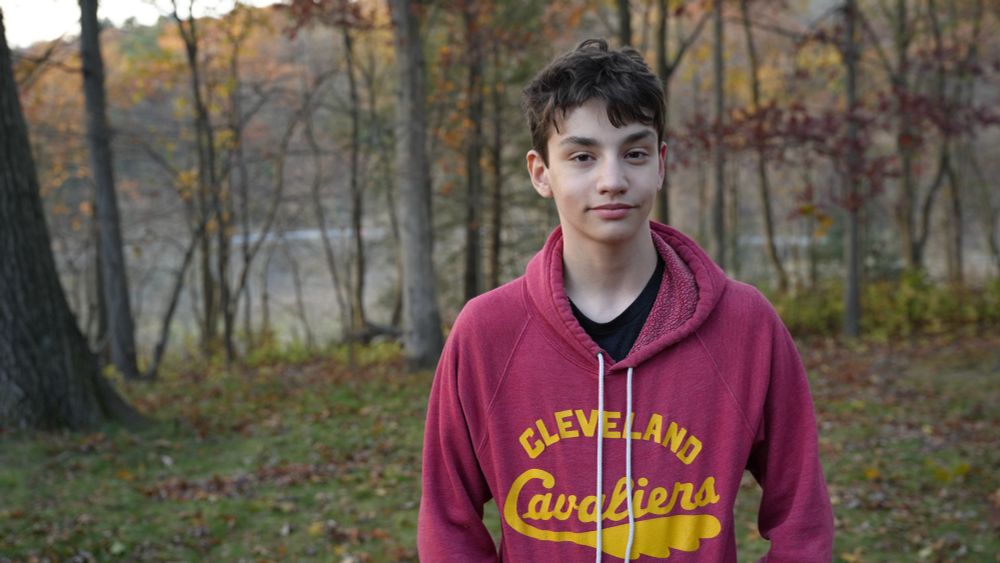 A boy, William Hidalgo Bowler, stands in front of a fall scene of trees. He is looking at the camera and smiling. He is outside, and it is daytime. 