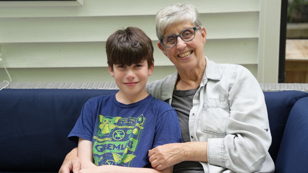  A child (Santiago Hidalgo-Bowler) and a woman (Jean Bowler) sit and smile on a blue couch together. Jean has her arm around Santiago’s shoulder. There is a white wall behind them. They are outside and it is daytime.