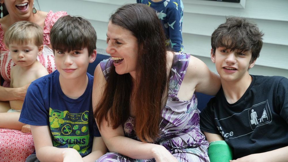 Two children (Santiago Hidalgo-Bowler and William Hidalgo-Bowler) and one woman (Alexandra Hidalgo) sit and smile together on a blue couch. Alex has her hand behind William and is looking at Santiago with a smile. There is a women and two children behind them, smiling. They are outside and it is daytime.