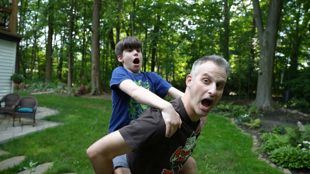 A man (Nathaniel Bowler) is standing in the grass and carrying a child (Santiago Hidalgo-Bowler) on his back. Santiago has his hands over Nate’s shoulders and is leaning back, an open-mouthed expression on both of their faces. There are trees behind them. They are outside and it is daytime.