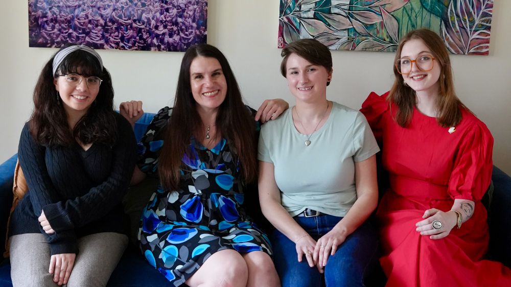 Four women (Natalie Cohen, Alexandra Hidalgo, Megan Simmermeyer, and Evelynn Pugh) smile and pose together on a blue couch. There are cream-colored walls behind them. It is daytime.