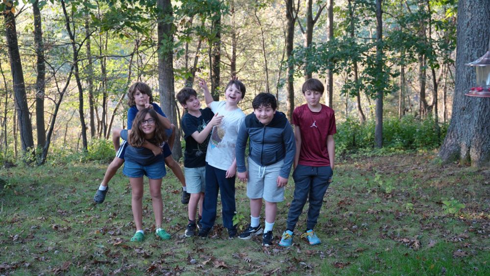 6 kids stand in front of trees and greenery. They pose for a picture, some making funny faces, some holding up peace signs, and one on their friends back. They are outside, and it is daytime.
