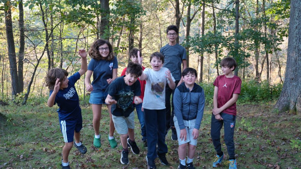 8 kids all pose for a photo. They are mid jump, frozen in the air. They are outside, and it is daytime. Behind them are trees and greenery.