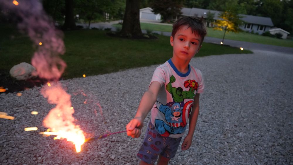 A child stands and holds a sparkler, lit up orange and pink, facing down on a driveway. He is looking at the sparkler inquisitively. There are trees and a house behind him. He is outside and it is nighttime.