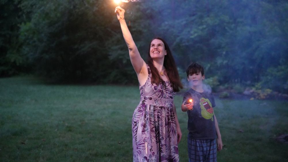 A woman, Alexandra Hidalgo, is holding a sparkler up above her head, and she is looking up at it. To her right, a child, Santiago Hidalgo Bowler, holds a sparkler out in front of him and looks off to the left. They are both outside, with grass and trees behind them. It is nighttime.