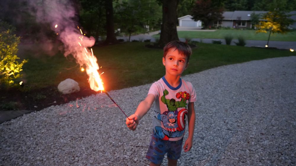 A child stands and holds a sparkler, lit up orange and pink, facing up on a driveway. He is looking at the sparkler inquisitively. There are trees and a house behind him. He is outside and it is nighttime.