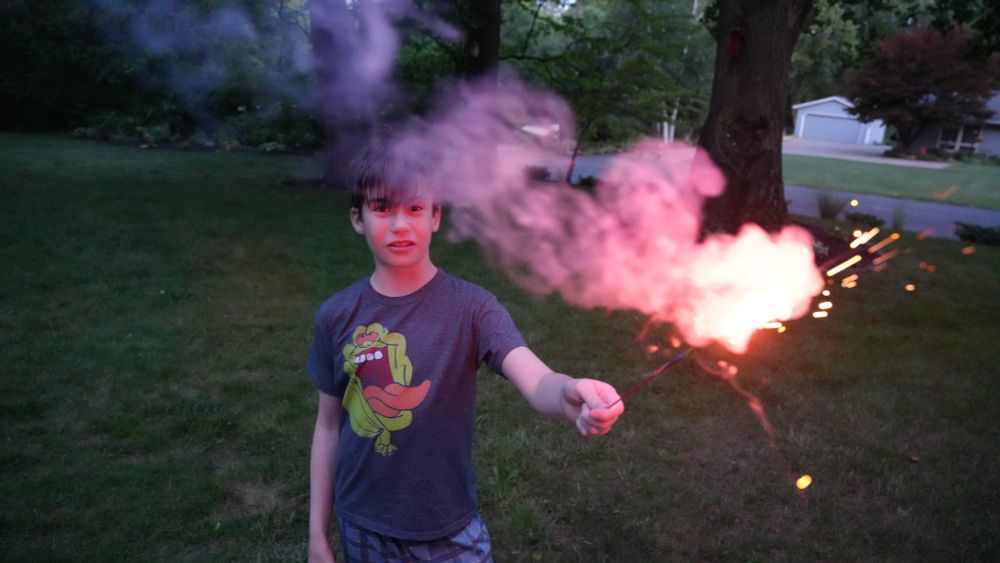 A child stands and holds a sparkler, lit up orange and pink, facing up in a field of grass. He is looking at the camera. There are trees, a road, and a house behind him. He is outside and it is nighttime.