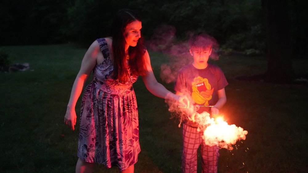 A woman, Alexandra Hidalgo, and a child, Santiago Hidalgo Bowler, look at the sparkler that the child is holding out in front of him. It is glowing orange and illuminating their faces and bodies. They are both outside, with grass and trees behind them. It is nighttime.