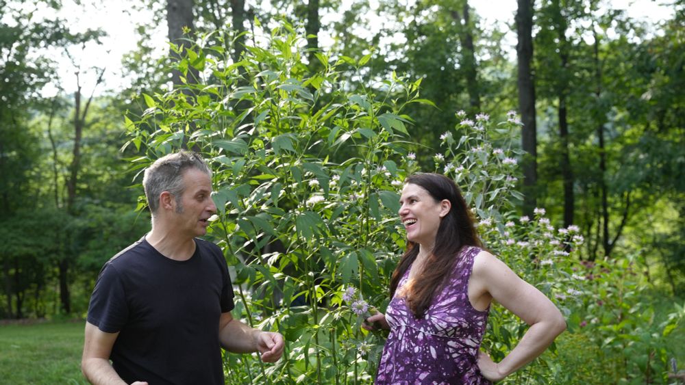 A man (Nathaniel Bowler) and a woman (Alexandra Hidalgo) stand and pose in front of a native garden. Nate is looking at Alex, mid-sentence, and Alex is looking at him with a smile. There are trees behind them. They are outside and it is daytime.