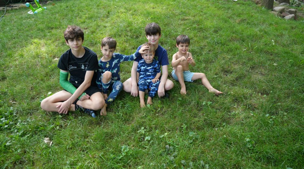 Five children (including William and Santiago Hidalgo-Bowler) sit and smile together in the grass. They are outside and it is daytime.
