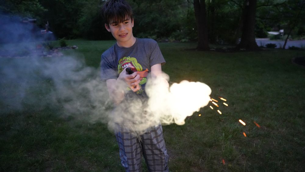 A child stands and holds a sparkler, lit up bright white, facing down in a field of grass. He is looking down with squinted eyes. There are trees behind him. He is outside and it is nighttime.