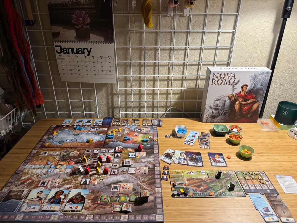 The board game Nova Roma set up on a bamboo desktop. The game is set up for solo and at an end game state with the automa scoring 91 and the sad human only scoring 69 (nice). There is lamp light directly over the game and behind the game is a wire grid wall with a January wall calendar visible and some tapestry yarn. 