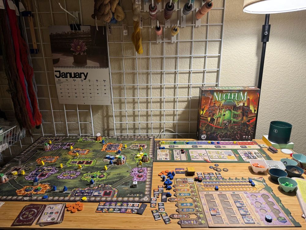 The board game Nucleum set up on a bamboo desktop for solo play at an end game state with lamp light from the right top corner. The game consists of a large main board with a green background and cities in orange and purple and green and white. There are yellow and blue meeples all over the board marking railways. To the right on the table is the player board that holds more meeples and card leads tiles for buildings and actions selection. Various other cardboard and wood pieces are scattered around and the milestone and scoring board is to the right and perpendicular to the main board. The box cover is visible in the back as well as a January wall calendar and some tapestry yarn on a grid wall behind the desktop. 