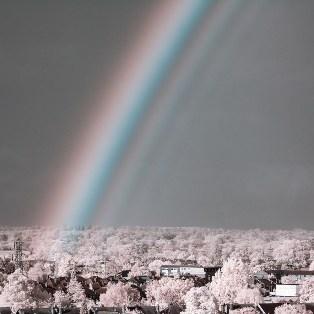 An infrared photo of a rainbow: a strange, ghostly-looking rainbow in a mostly black and white photo, with a faintly red outer edge and a cyan inner edge, and what look like a few even fainter ‘echo’ bows inside
