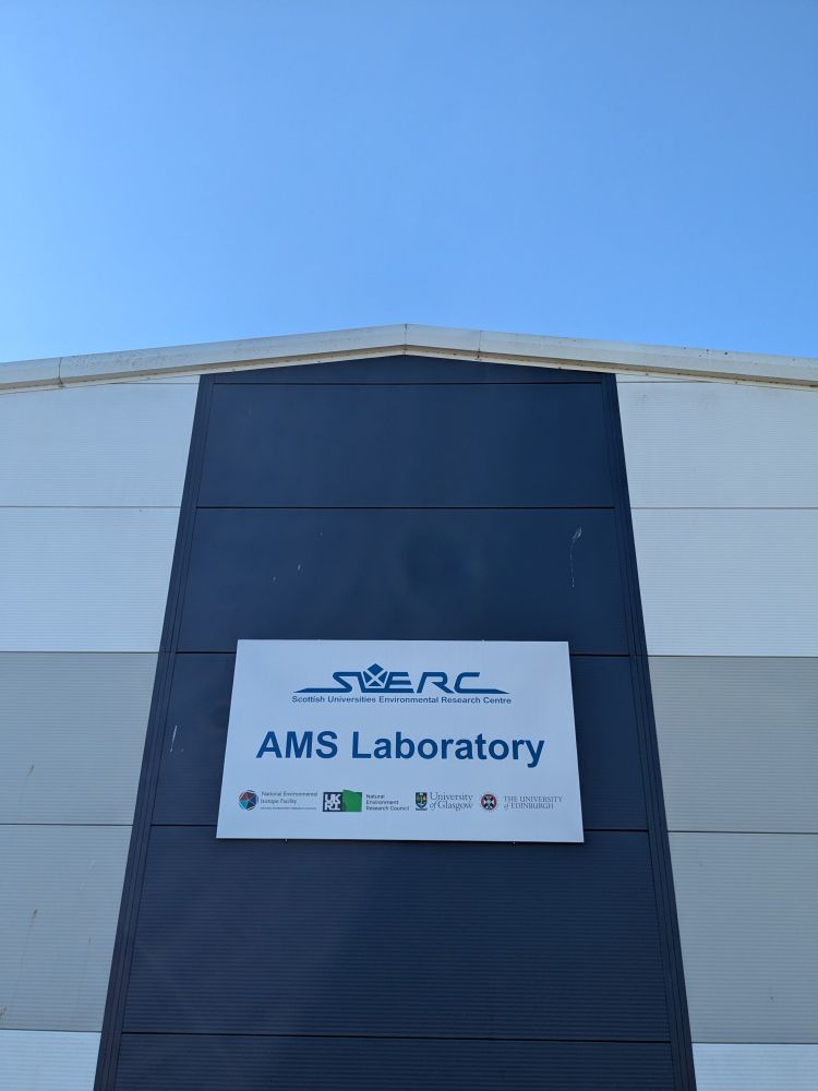 A photo of the side of the AMS Laboratory at the Scottish Universities Environmental Research Centre (SUERC) with lovely clear blue skies above it. A sign on the side of the building includes the logo for SUERC, "AMS Laboratory", and different universities and organisations associated with it. 