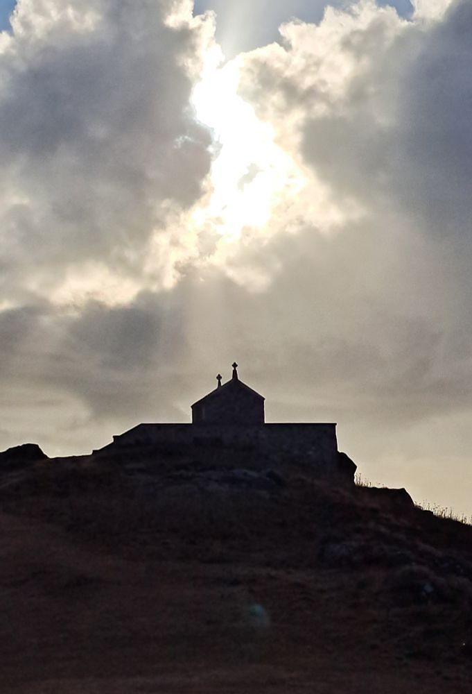 A silhouetted chapel on a hill with moody sky and sunburst behind cloud