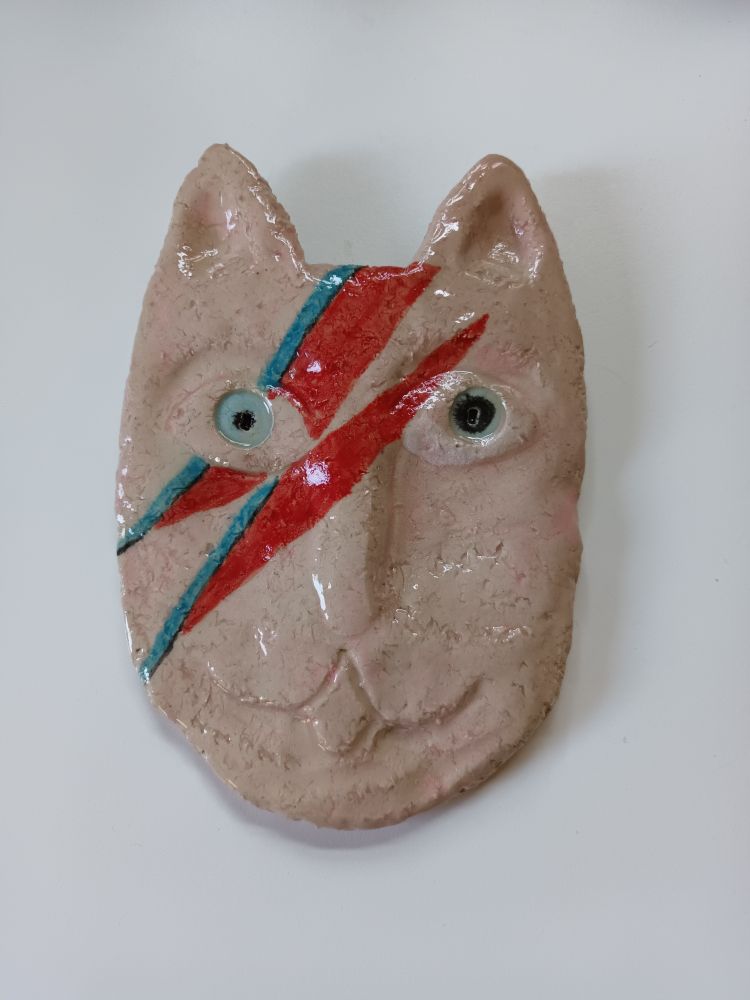 A pottery wall plaque of a cats head but decorated like David Bowie's Aladdin Sane album cover with a lightning streak.