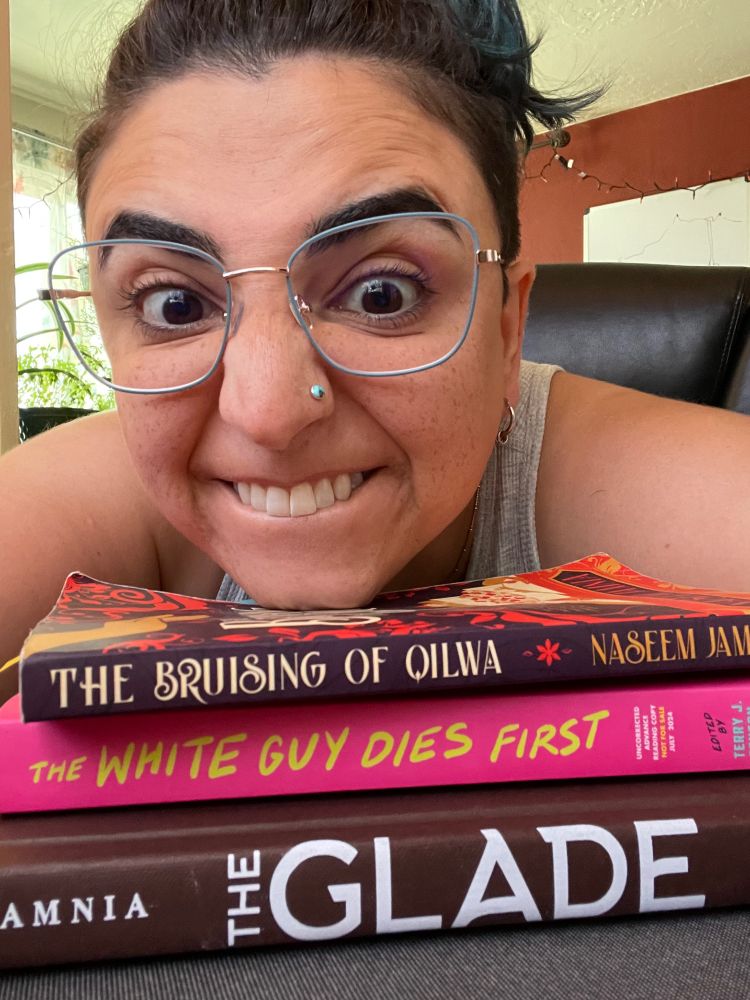 Naseem, a light-skinned West Asian person with brown hair pulled back, blue-rimmed glasses, and a turquoise nose stud, props their chin on a stack of their books: The Bruising of Qilwa, The White Guy Dies First, and The Glade. They're baring their upper teeth in a silly expression.