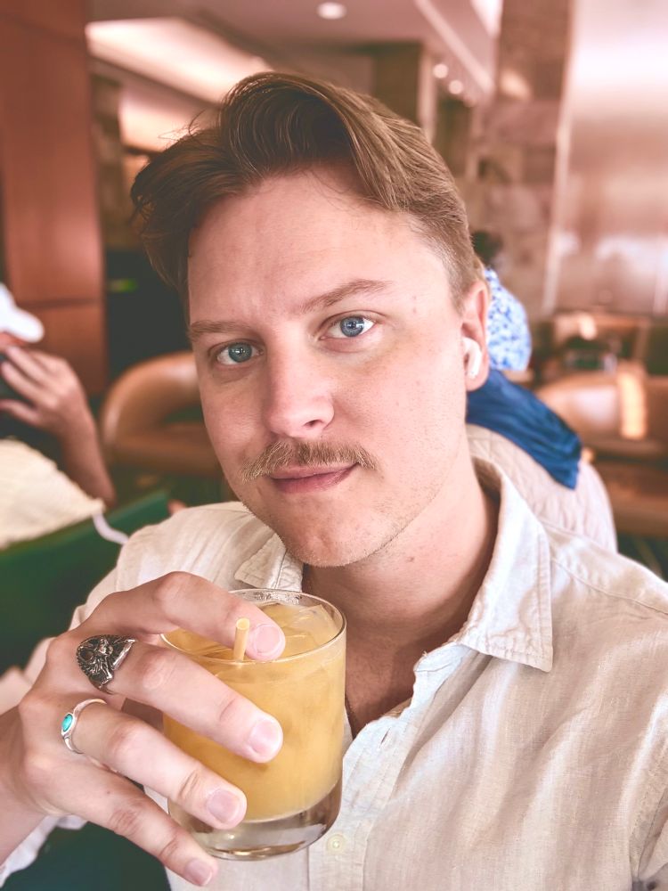 Photo of Jacob Thomas raising a glass with orange liquid in it toward the camera. 