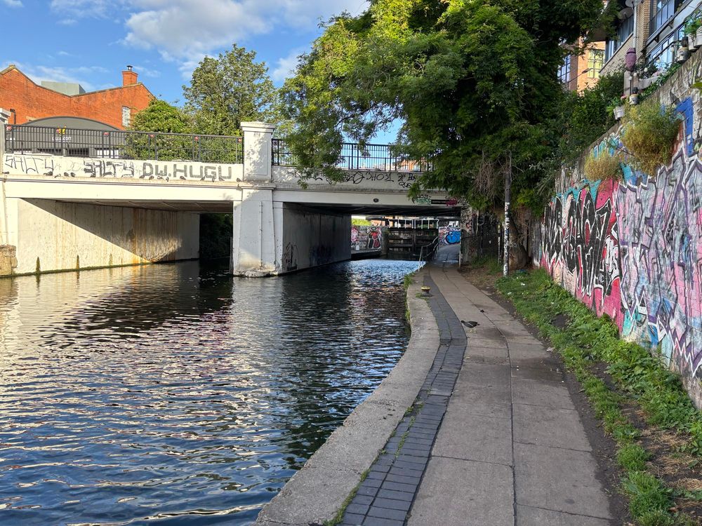 A section of the Regent’s Canal looking west towards the Kentish Town Road Bridge. There is a moorhen on the towpath, and a lock gate is visible under the bridge.