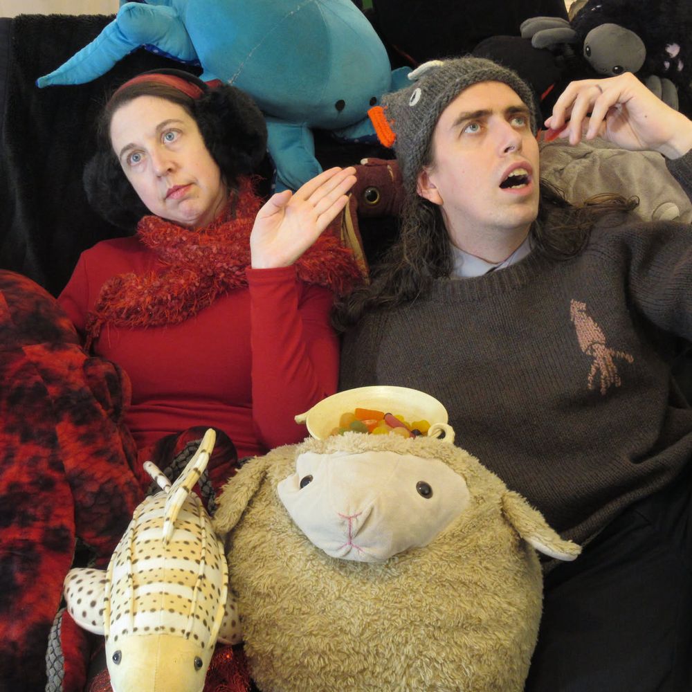 A photo of a white woman in a red shirt wearing enourmous black earmuffs and a white man with long brown hair wearing a knitted hat shaped like a fish and a brown sweater with a squid image.  They are frozen in positions reminiscent of people in medieval/renaissance paintings.  The man has his mouth open in a fish like manner and is poised to eat a gummy fish candy.  The woman has her hand palm out  to the side and is looking utterly devoid of emotion.  They are surrounded by a plethora of plush animals including a sheep, a fish, a spider, and several octopus.  The sheep is central and has a bowl of gummy candies on it's head.