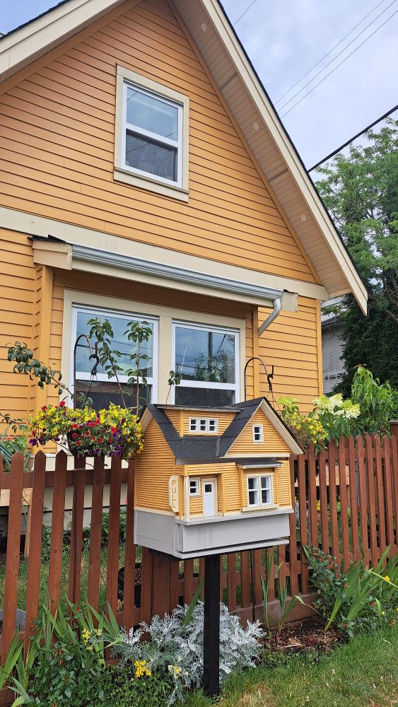 A yellow house has a small replica of itself in front of it; small house is a little free library 