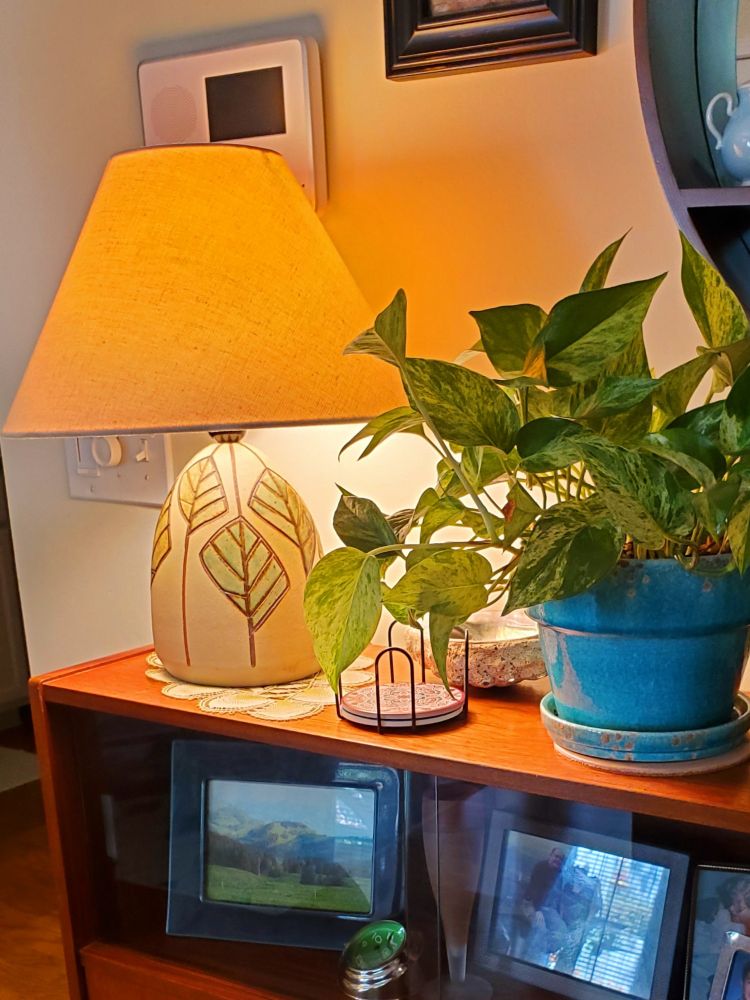 A 1960s era lamp sits on a wooden sideboard. The lamp is egg-shaped, with large, simplified leaf designs carved into the rough cream texture of the lamp. The leaves are glazed in colors ranging from gold to turquoise. The lamp is on, a gold light illuminating a spreading pothos plant in a turquoise pot next to it.