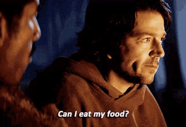 GIF of Cassian Andor (played by Diego Luna) cast in firelight, wearing a poncho. He turns his head and asks, mouth full, "Can I eat my food?"