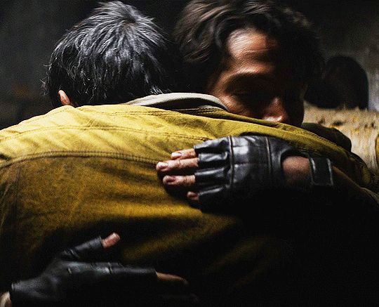 A .GIF of Cassian Andor wrapping his arms around Brasso in episode 12, season 1 of ANDOR. Brasso is wearing a golden, worn-looking coat that catches a beam of light across his shoulders. The same light falls onto Cassian's forehead and the bridge of his nose, buried into his friend's shoulder in his grief. The background of the shot is dark as the two figures are meeting in the sewers. It is the type of hug you need when you've lost someone, and feel unmoored.