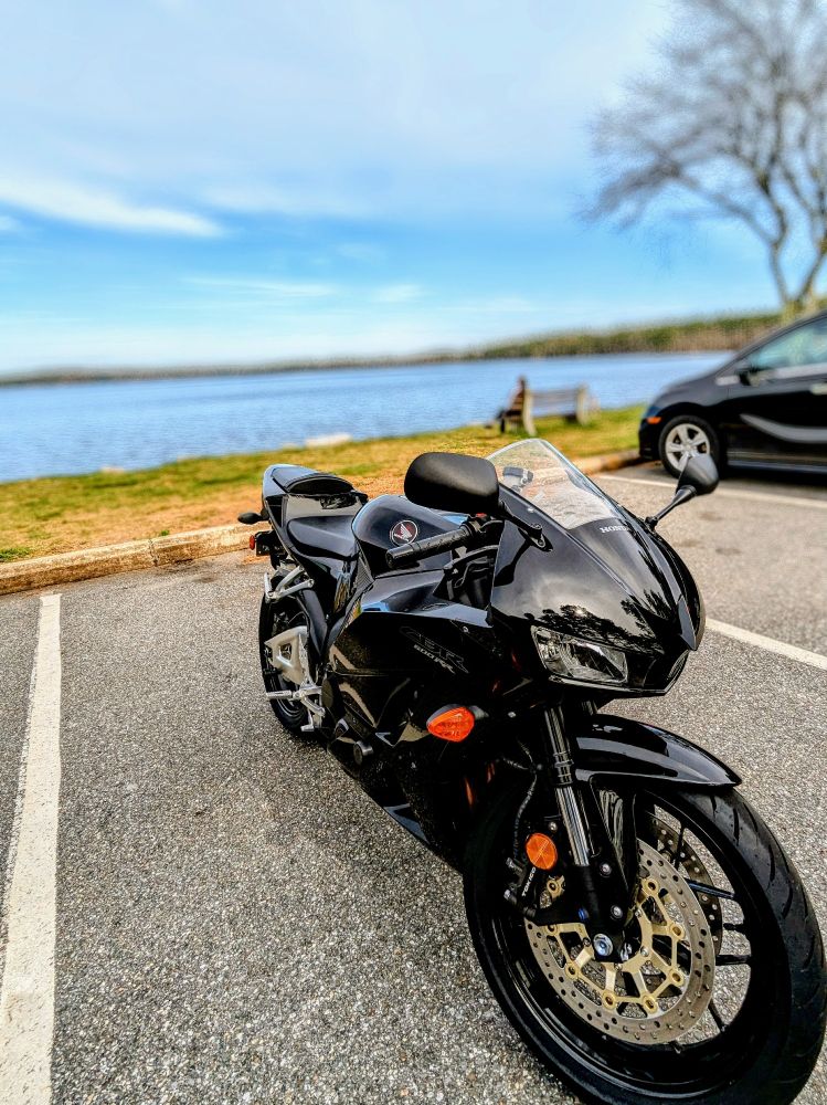 Shiny black motorcycle (2014 Honda CBR600RR) parked next to a lake, with blue skies and wispy clouds overhead.