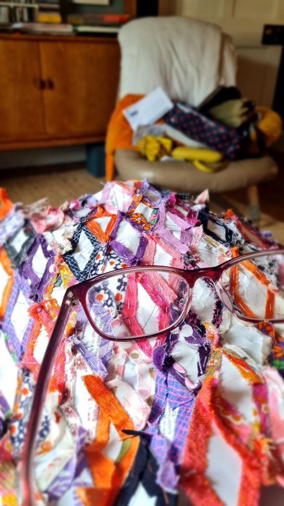 Photo of the reverse side of my current patchwork project sitting on my knee, with my reading glasses sitting on top. The pieces in the patchwork are diamond shaped and have been chosen from my cotton fabric scrap bag. They are mainly pink, purple, orange and navy. They have been sewn together by hand over many hours. in the background is a chair with bags, cushions, a quilt and a letter piled up on it.