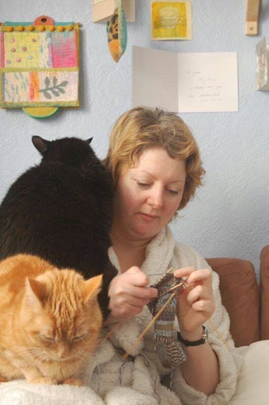 A 40 year old woman wearing a white dressing gown sits knitting a sock on 4 needles while a black cat perches on her shoulder and a ginger cat sits on her leg. The cats are impeding the knitting, but she seems intent on carrying on.