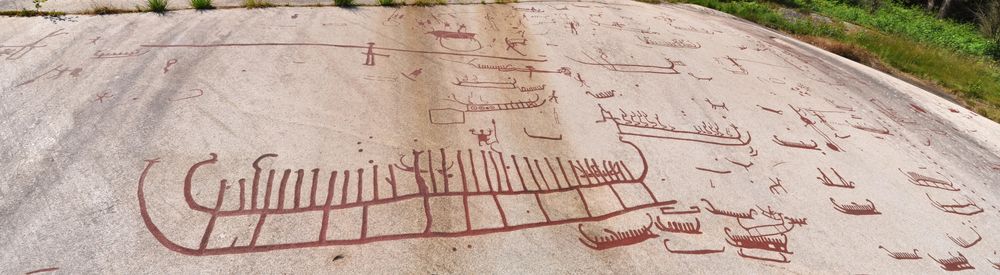 Stitched panorama of the main panel of engravings at Tanum, Sweden. It shows many boats with upturned bows and lines above the hull which are presumably stylised people. There is an image of one big boat in the centre and many smaller boats around it. Following Swedish practice, the engravings are picked out in red paint so they can be seen without observers making the lines deeper so they can take a nice photo.