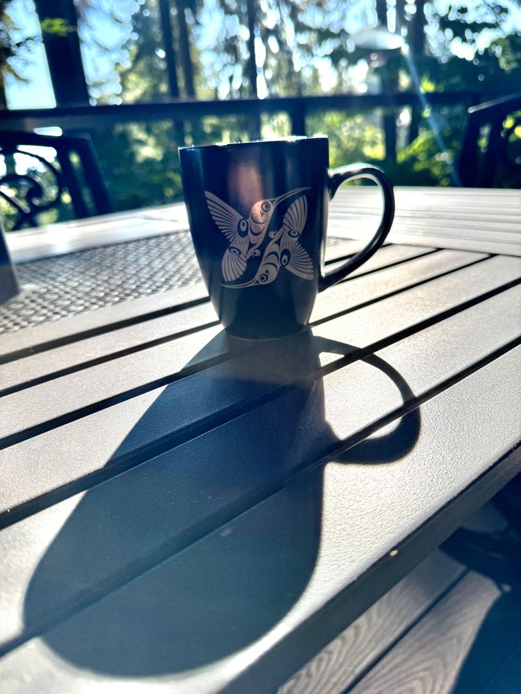 A black coffee mug painted with an Indigenous Pacific Northwest artistic rendering of two hummingbirds in flight, depicted in light gray. The mug is resting on a dark metal table and a long shadow of the mug stretches toward the viewer, the result of the morning sun positioned just above the edge of the red umbrella visible in the top of the frame. Out of focus Douglas fir trees behind a deck rail populate the background 