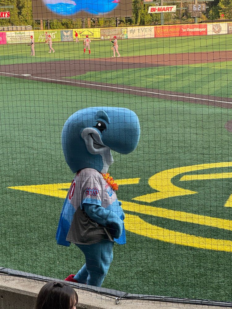 Photo taken from seats behind home plate at a baseball game, a mascot wearing a stylized whale costume, a baseball jersey and a lei is walking on the field