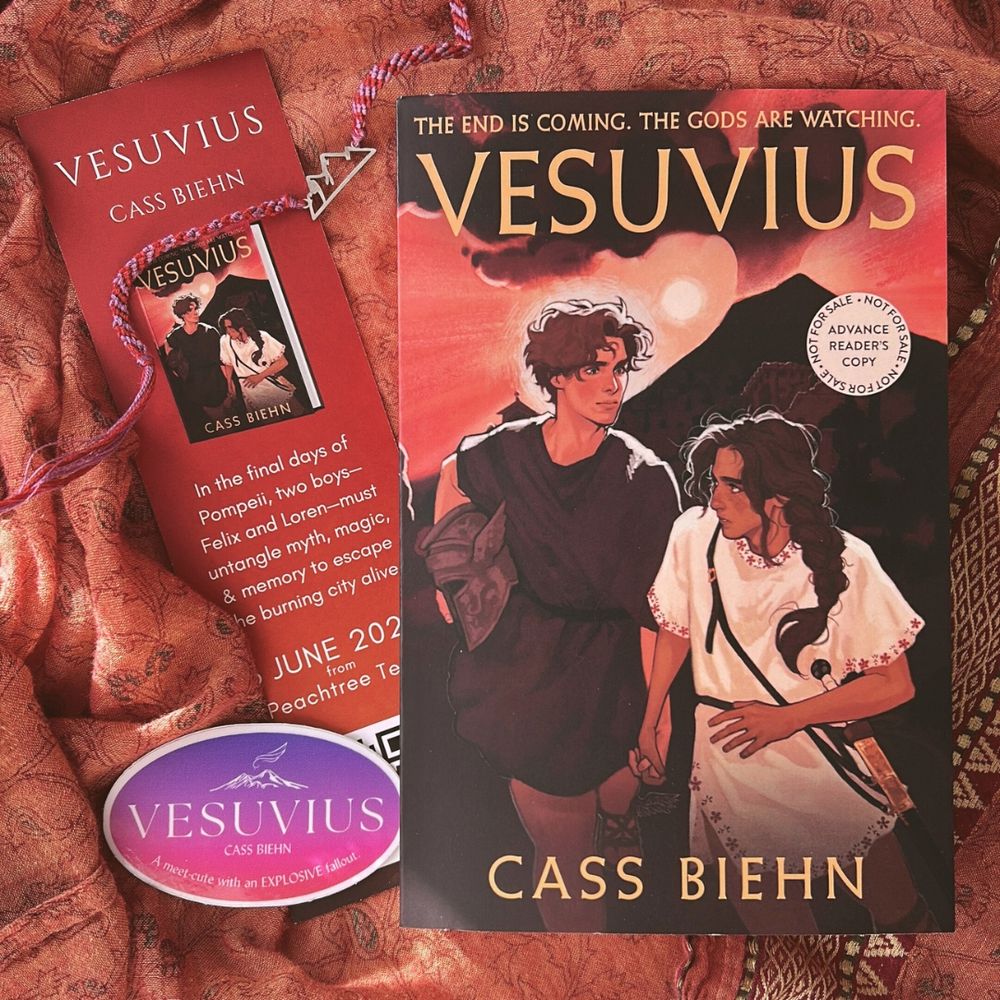 orange/gold textured fabric background with a paperback ARC of Vesuvius surrounded by a purple/pink sticker, a bookmark that matches the cover, and a purple/red bracelet with a mountain charm