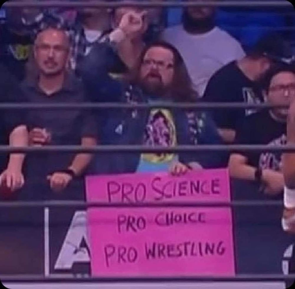 A man in a crowd holding a sign at an AEW event that says “Pro science, pro choice, pro wrestling”