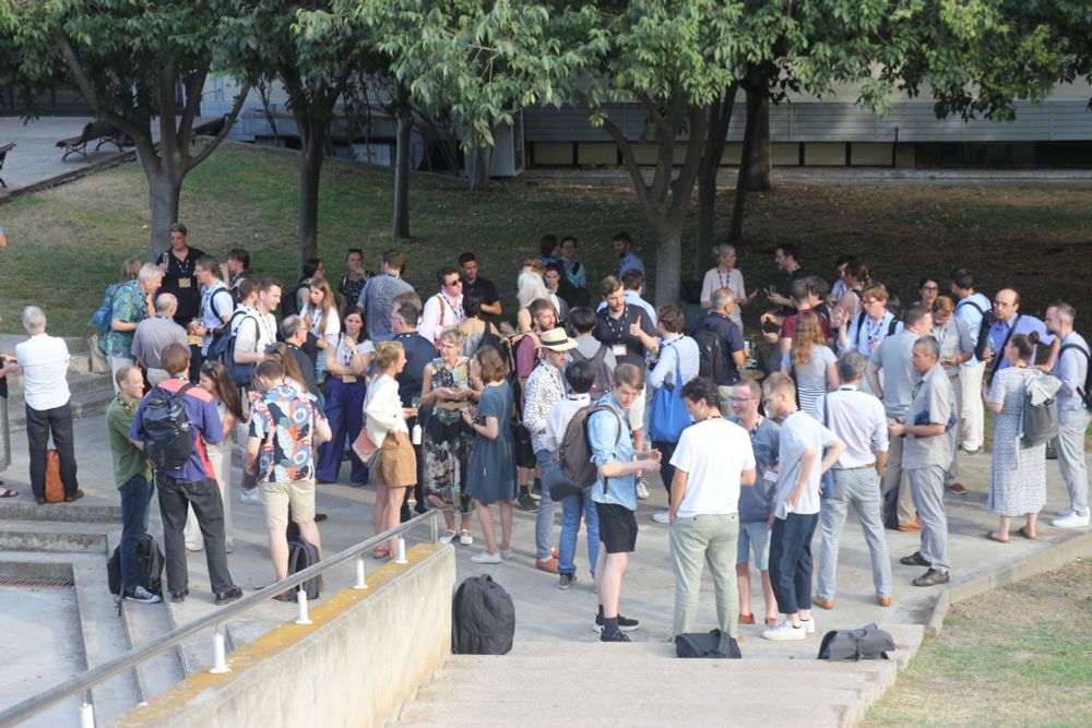 A group of political scientists are standing under the cover of trees at a wine reception on a concrete concourse