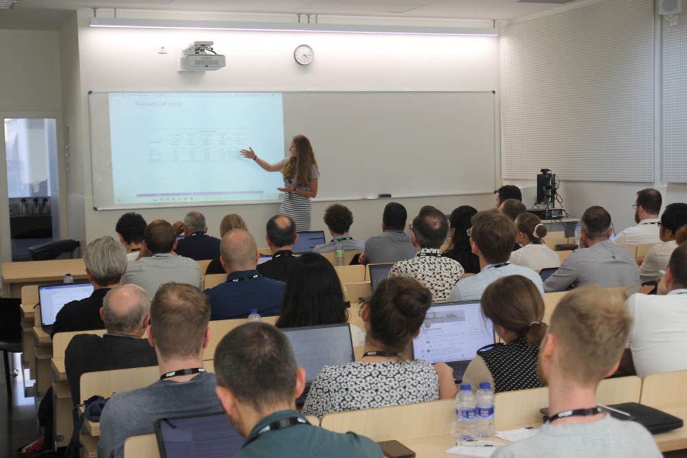 A presenter is pointing at slides while giving an overview of her research, with lots of people in the audience sitting in rows to listen and make notes