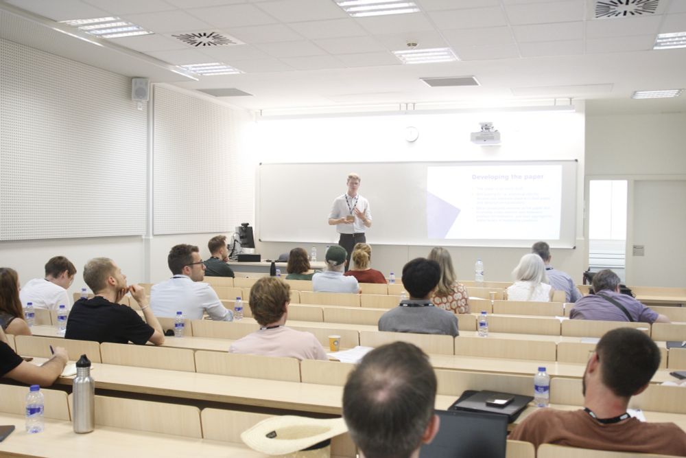 Conference presentation with rows in the foreground with people listening to a presenter towards the back of the picture